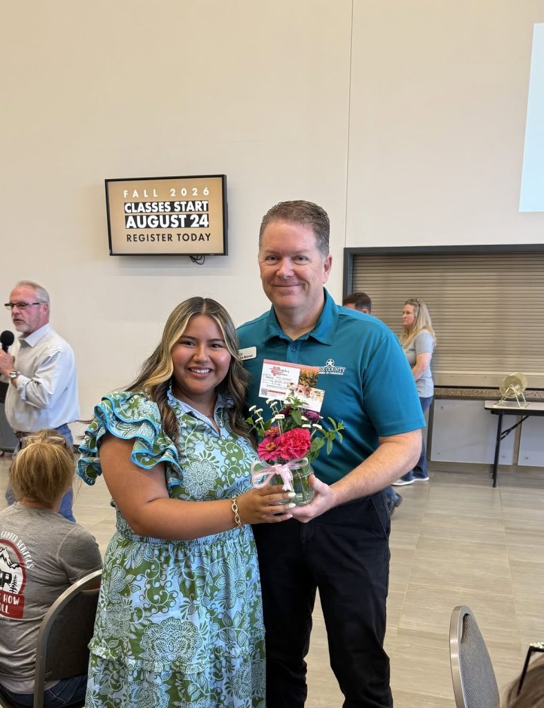 A Wise County Chamber of Commerce representative presents flowers to a community member during a recognition ceremony, with a "Fall 2026 Classes Start" sign visible in the background